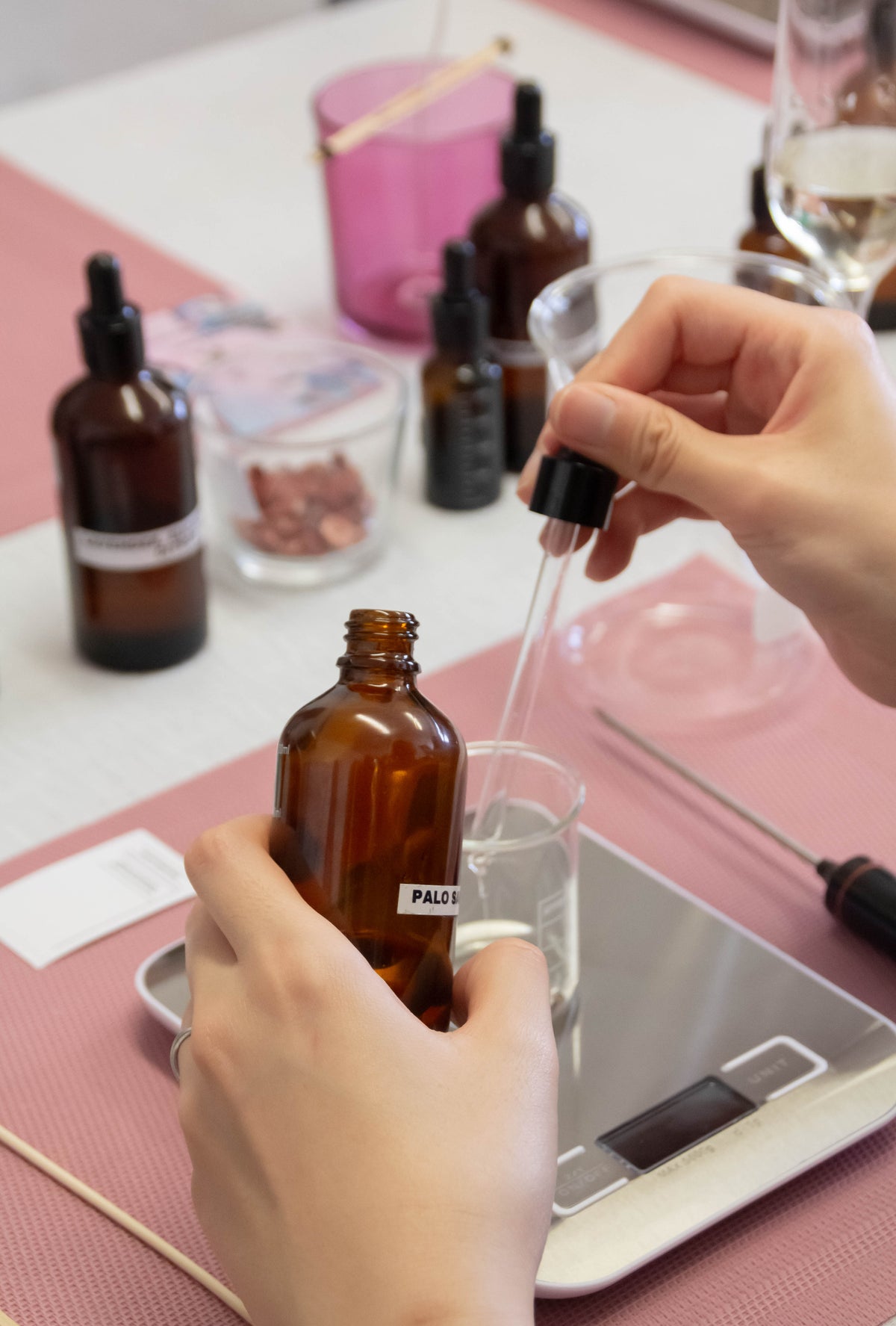 A close-up of hands preparing an amber glass dropper bottle of fragrance oils on a pink and white tabletop. One hand holds the bottle while the other uses a glass dropper to transfer liquid into a small clear container on a digital scale.