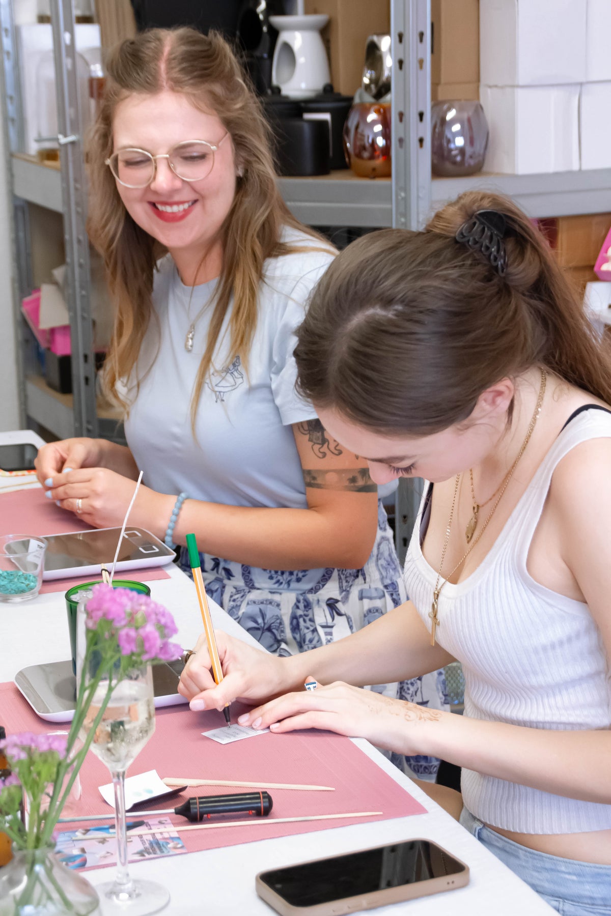 Two women sitting at a table in a craft or workshop setting, working on small projects with pink mats, tools, and flowers in the foreground.