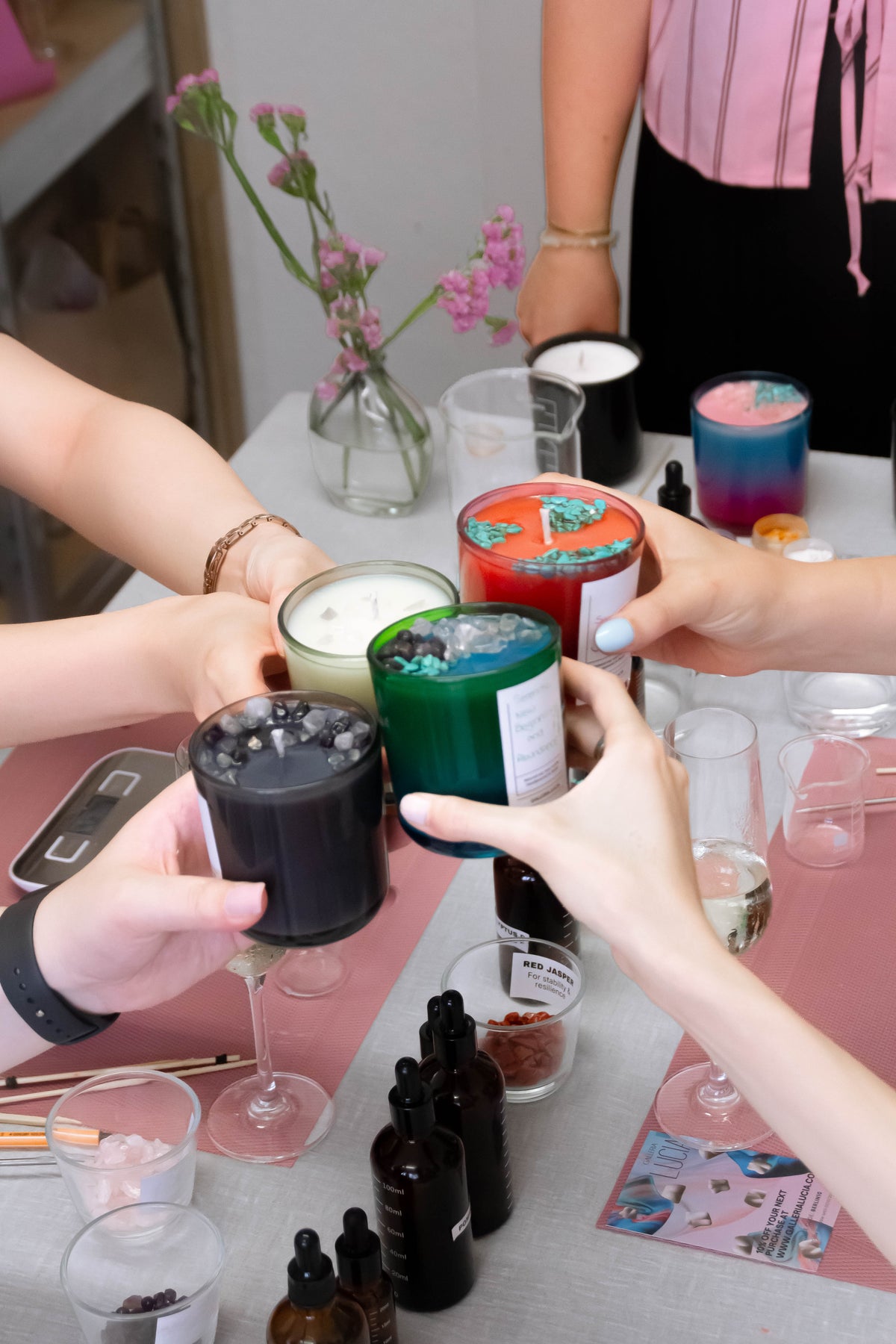 Several hands hold colorful soy candles in colourful glasses, clinking together on a table with small bottles, cups, and flowers in the background.