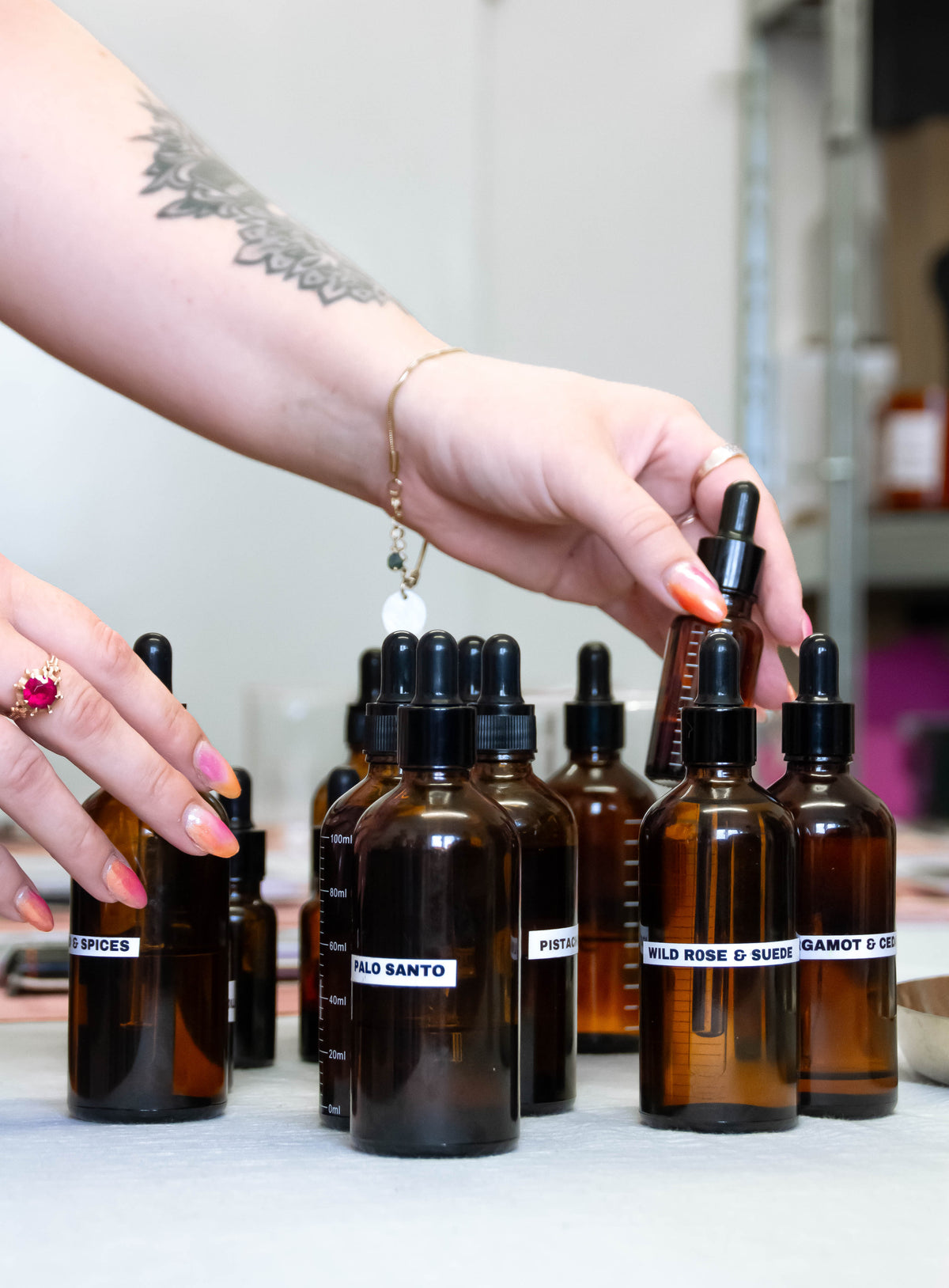 A close-up of hands arranging amber glass dropper bottles with fragrance oils on a light surface. The bottles have white labels with handwritten text such as 'PALO SANTO' and 'WILD ROSE'.