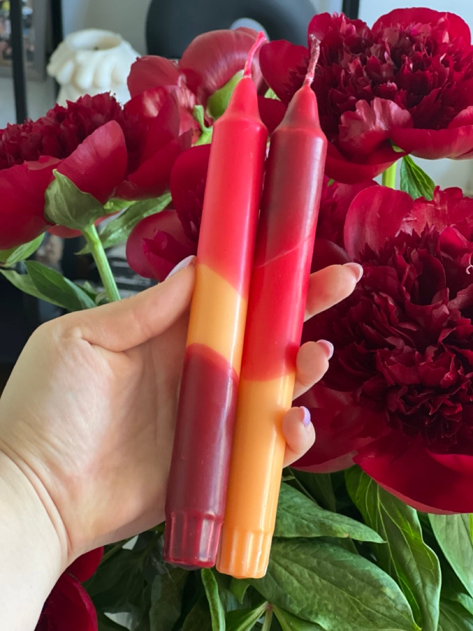 Hand holding two red and orange gradient candles in front of red flowers