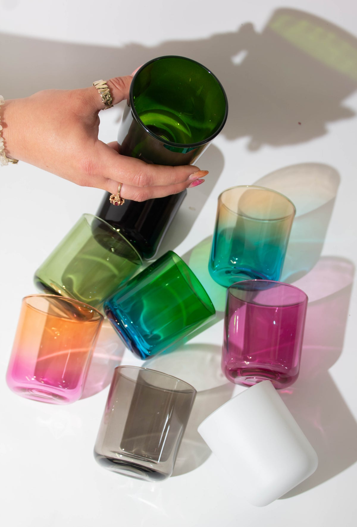 A hand holds a dark green candle jar above a group of colorful candle jars on a white surface, including green, blue, pink, orange, gray, and white.