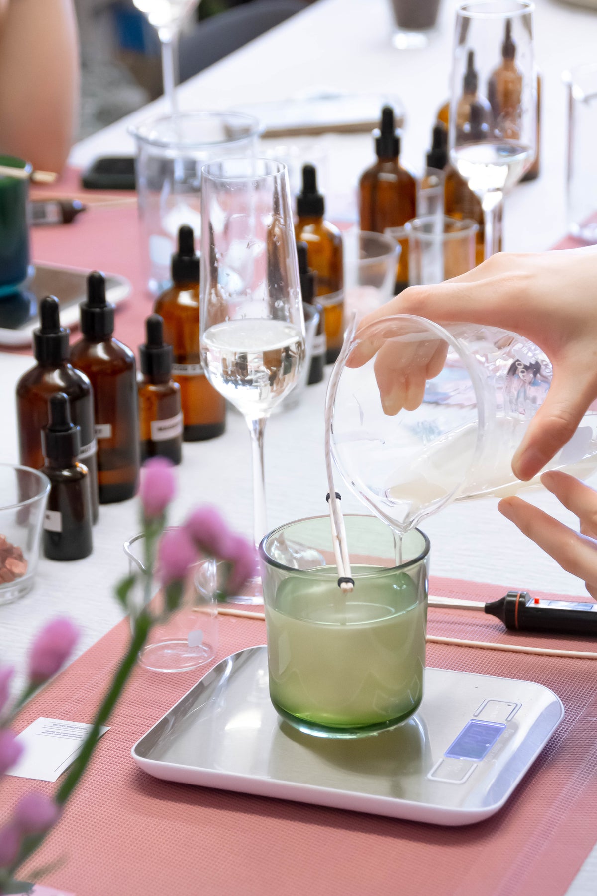 A hand pours liquid wax from a glass beaker into a green glass container placed on a digital scale, set on a pink table runner with fragrance bottles in the background.